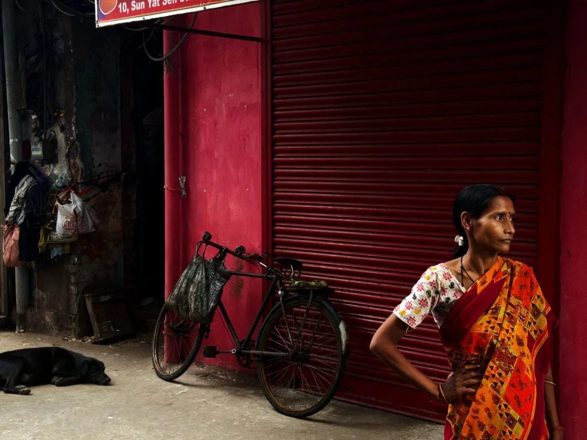 Leica Lux 2.0’a Steve McCurry İmzalı Yeni Görünüm Geldi 23 A woman in a patterned sari stands before a closed red metal shutter, with a bicycle parked beside her and a black dog resting on the ground.