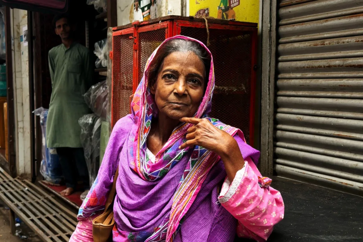 Leica Lux 2.0’a Steve McCurry İmzalı Yeni Görünüm Geldi 24 An elderly woman wearing a pink and purple patterned headscarf and matching tunic stands in front of a red metal cage, with a man in a green kurta visible in the background.