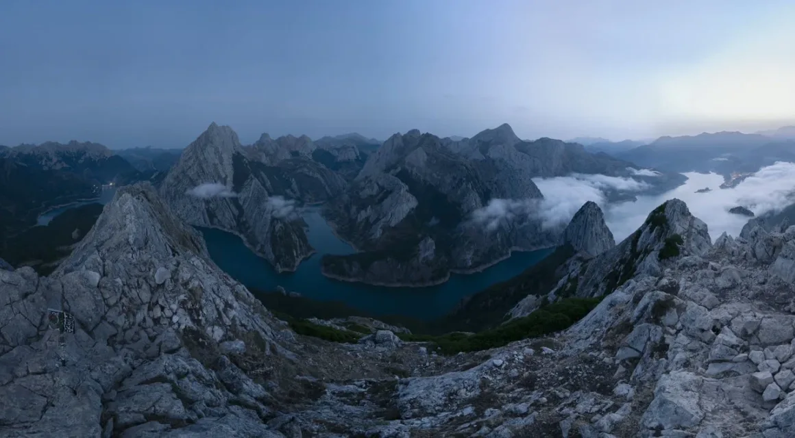 Angel Fux, Tam Güneş Tutulmasını Tek Bir Fotoğrafta Zamana Dönüştürdü 25 A panoramic view of a deep blue mountain lake winding through jagged, rocky peaks under a twilight sky, with low-hanging clouds.