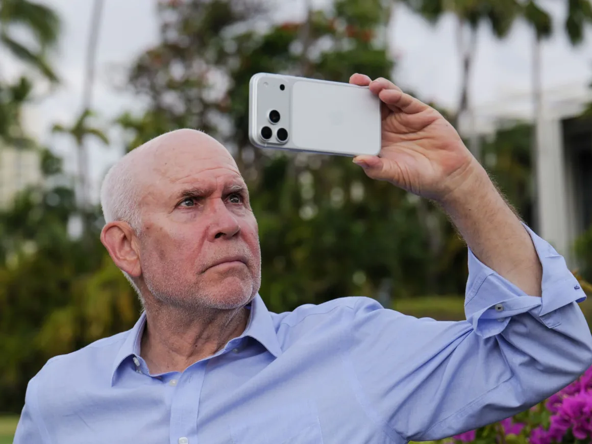 Leica Lux 2.0’a Steve McCurry İmzalı Yeni Görünüm Geldi 20 Başı tıraşlı, açık mavi düğmeli gömlek giyen yaşlı bir adam açık havada beyaz bir akıllı telefonla fotoğraf çekiyor.