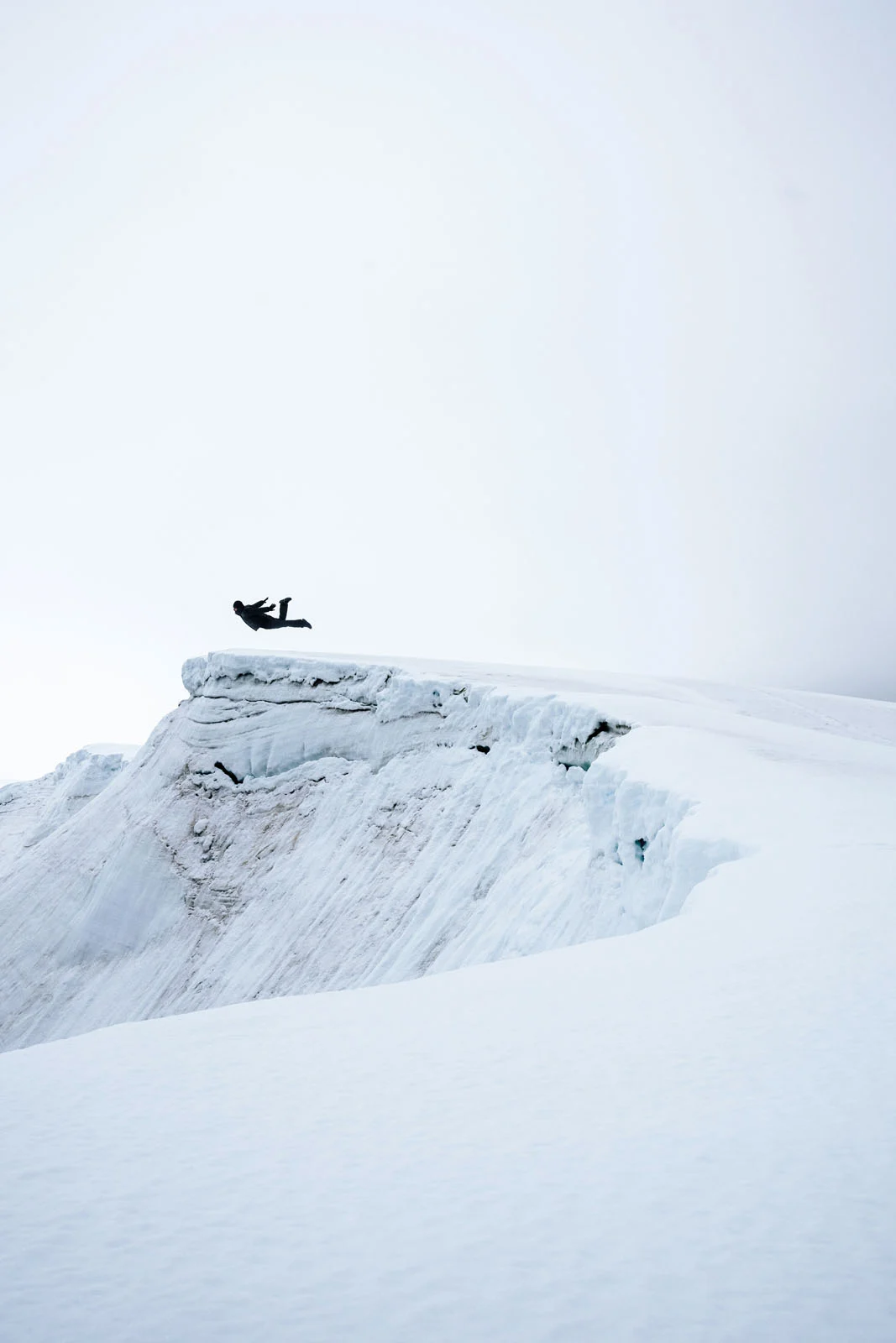A person in dark clothing leaps horizontally through the air above a snow-covered ridge.