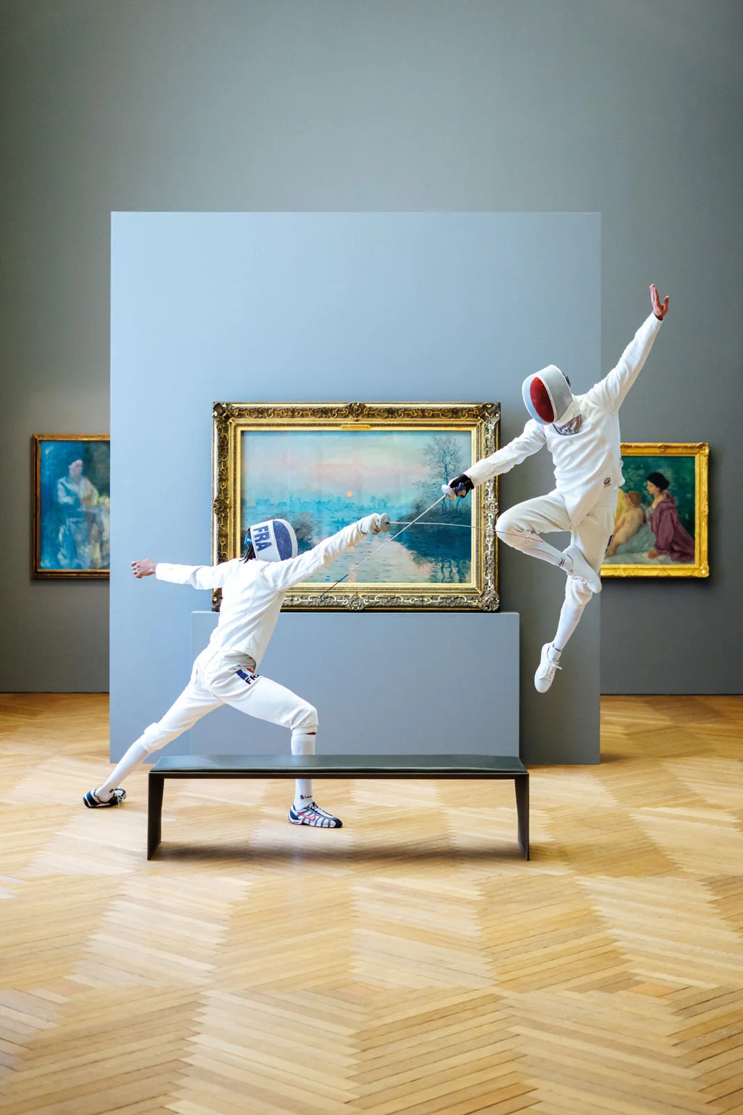 Two fencers in white uniforms and masks duel in a museum gallery, with one lunging from a bench and the other leaping mid-air.
