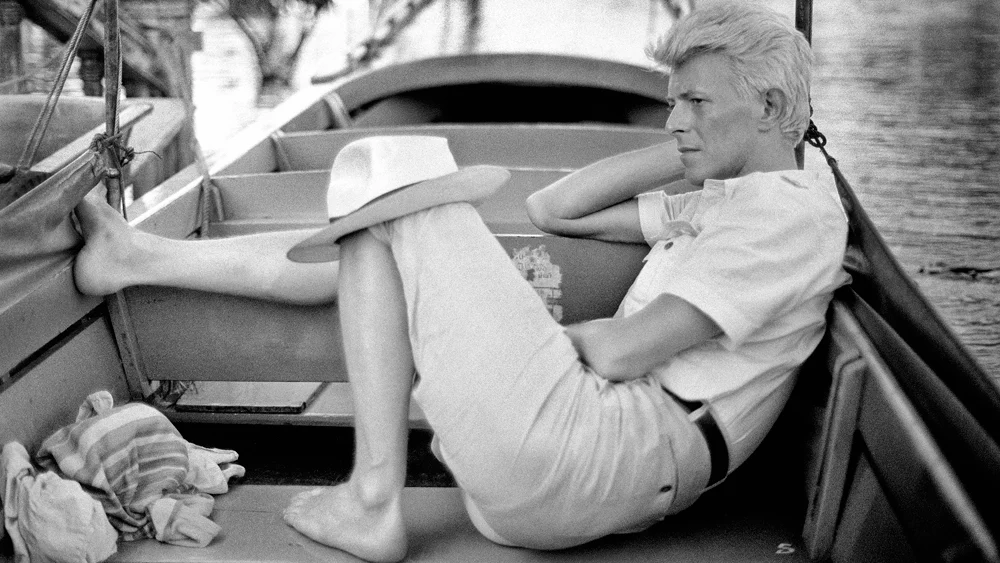 David Bowie Fotoğrafları Sürükleyici Bir Deneyime Dönüşüyor 20 David Bowie, Bangkok’ta açık renk gömlek ve pantolonla ahşap teknede çıplak ayakla oturuyor; şapkasını dizinin üzerinde tutuyor.