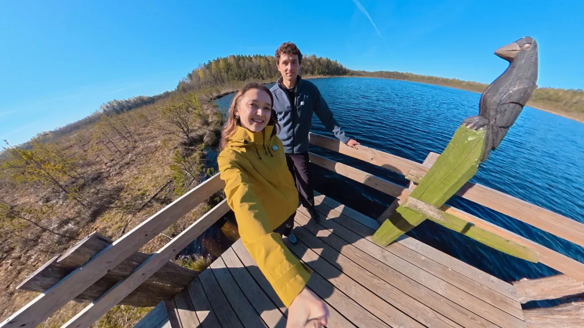 A young woman and man take a selfie on a wooden observation deck overlooking a lake and marshland, with a carved wooden bird sculpture nearby.