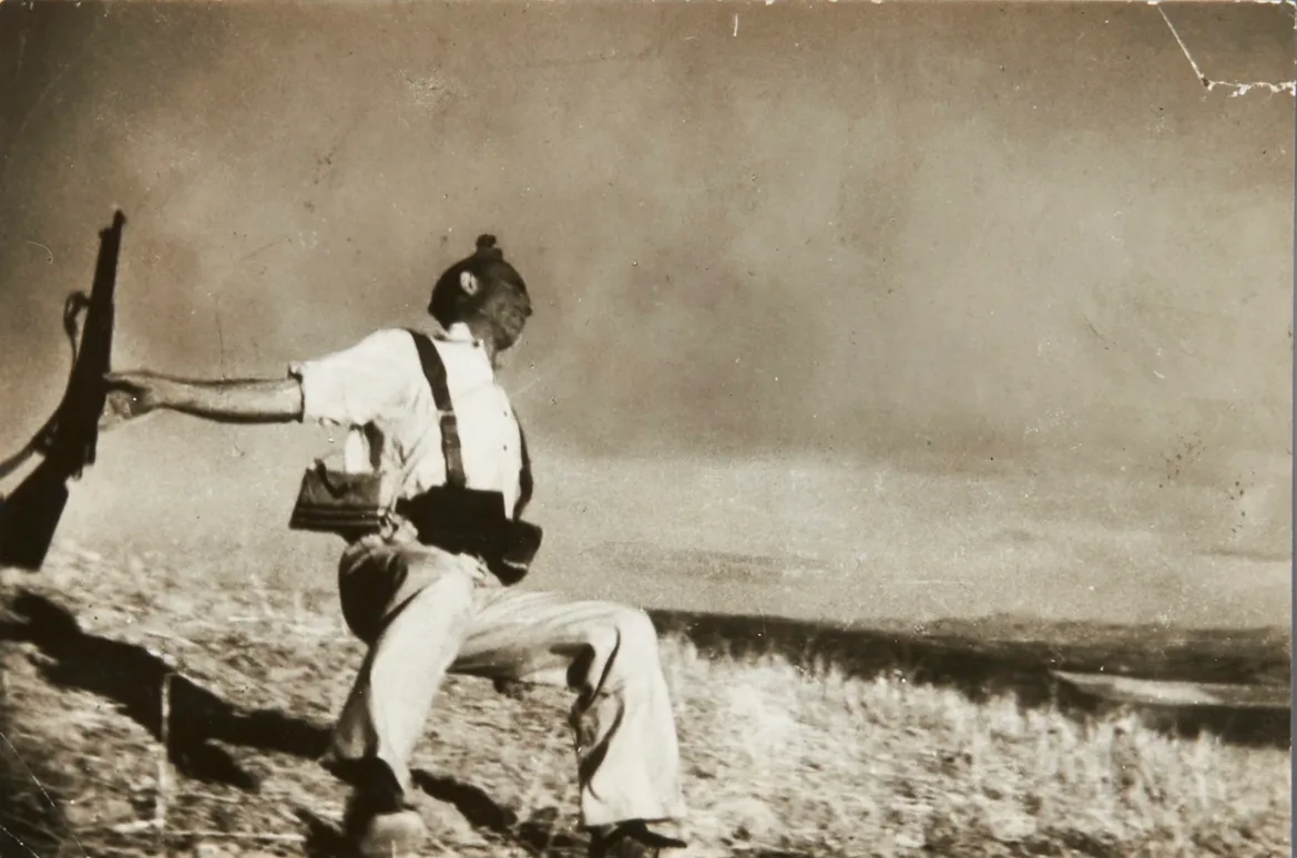 A sepia-toned photograph shows a soldier in a white shirt and suspenders falling backward on a grassy hillside while holding a rifle in his outstretched hand.