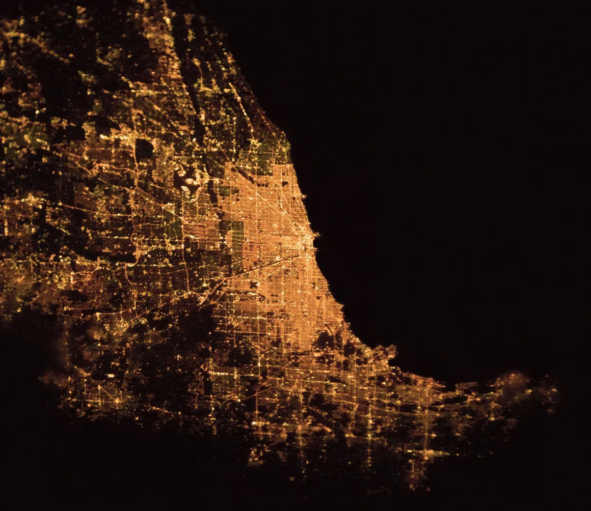 Astronotun Önce-Sonra Fotoğrafları Kentlerin Turuncudan Beyaz Işığa Geçişini Gösteriyor 19 A satellite view of a brightly lit urban area at night, showing a dense grid of city lights along a dark coastline.