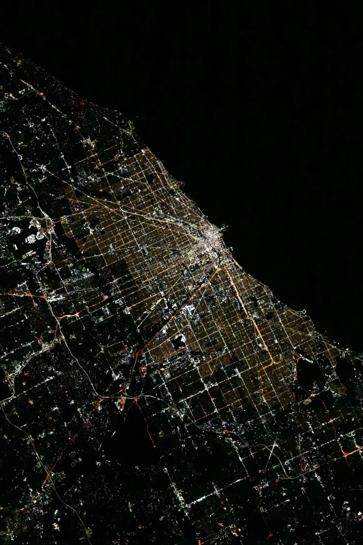 Astronotun Önce-Sonra Fotoğrafları Kentlerin Turuncudan Beyaz Işığa Geçişini Gösteriyor 20 An aerial night view of a brightly lit city grid along a dark coastline, with major roads and highways forming distinct patterns.