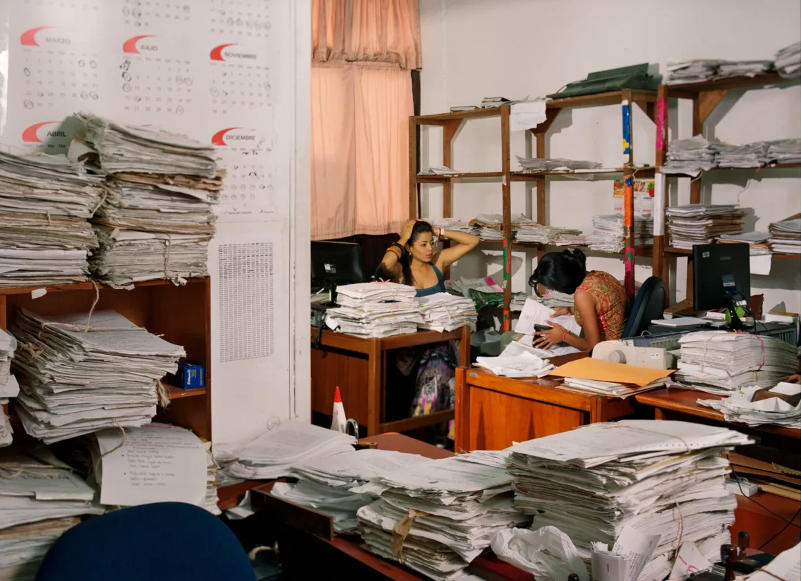 Dünyanın Dört Bir Yanından Bürokrat Portreleri 23 İki kadın, büyük kâğıt yığınları ve dosyalarla dolu rafların bulunduğu dağınık ofiste masalarında çalışıyor.