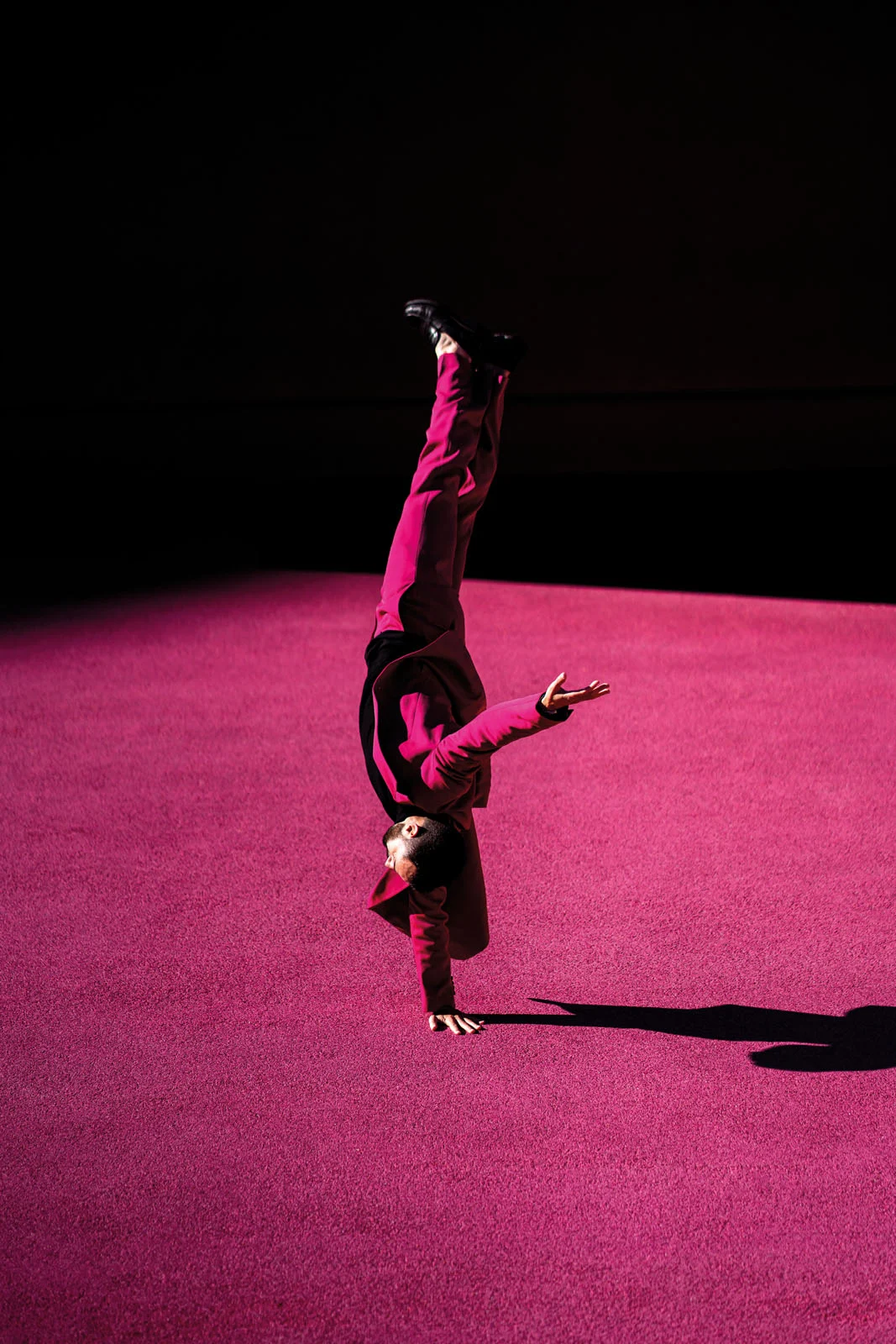 A person in a bright pink suit performs a one-handed handstand on a vibrant pink surface against a dark background.