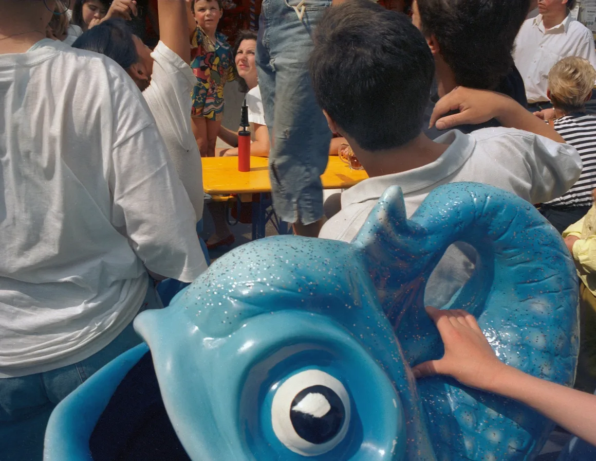 Wolfgang Zurborn’un Kalabalıkları: Toplum Değişmeden Önce 1980’ler ve 1990’lar 27 Öndeki parlak mavi denizatı biçimli lunapark koltuğunun arkasında insanlar toplanıyor.