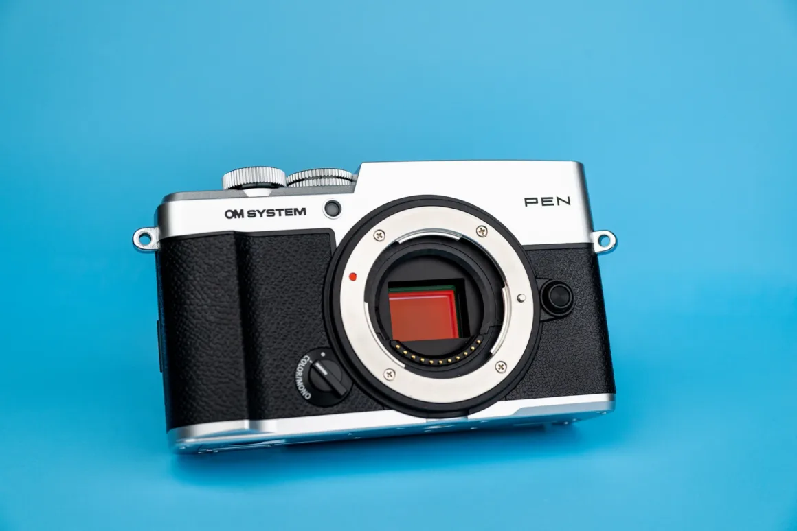 OM System PEN İncelemesi: Tanıdık Ama Son Derece Şık 23 A silver and black OM SYSTEM PEN mirrorless camera body with an exposed sensor, set against a solid blue background.