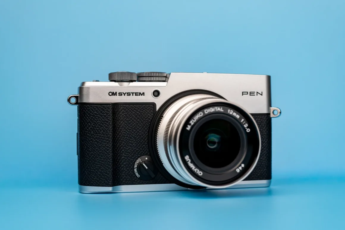 OM System PEN İncelemesi: Tanıdık Ama Son Derece Şık 18 A silver and black OM SYSTEM PEN digital camera with a lens attached, centered against a solid light blue background.
