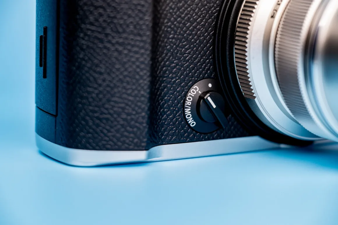 OM System PEN İncelemesi: Tanıdık Ama Son Derece Şık 29 A close-up of a camera body featuring a physical switch labeled "COLOR/MONO" set to the color position, with a silver lens visible on the right against a light blue background.