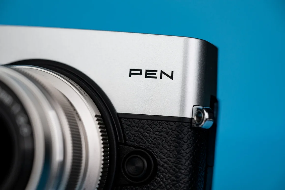 OM System PEN İncelemesi: Tanıdık Ama Son Derece Şık 19 A close-up view of a silver and black camera body with the word "PEN" printed on the top panel against a solid blue background.