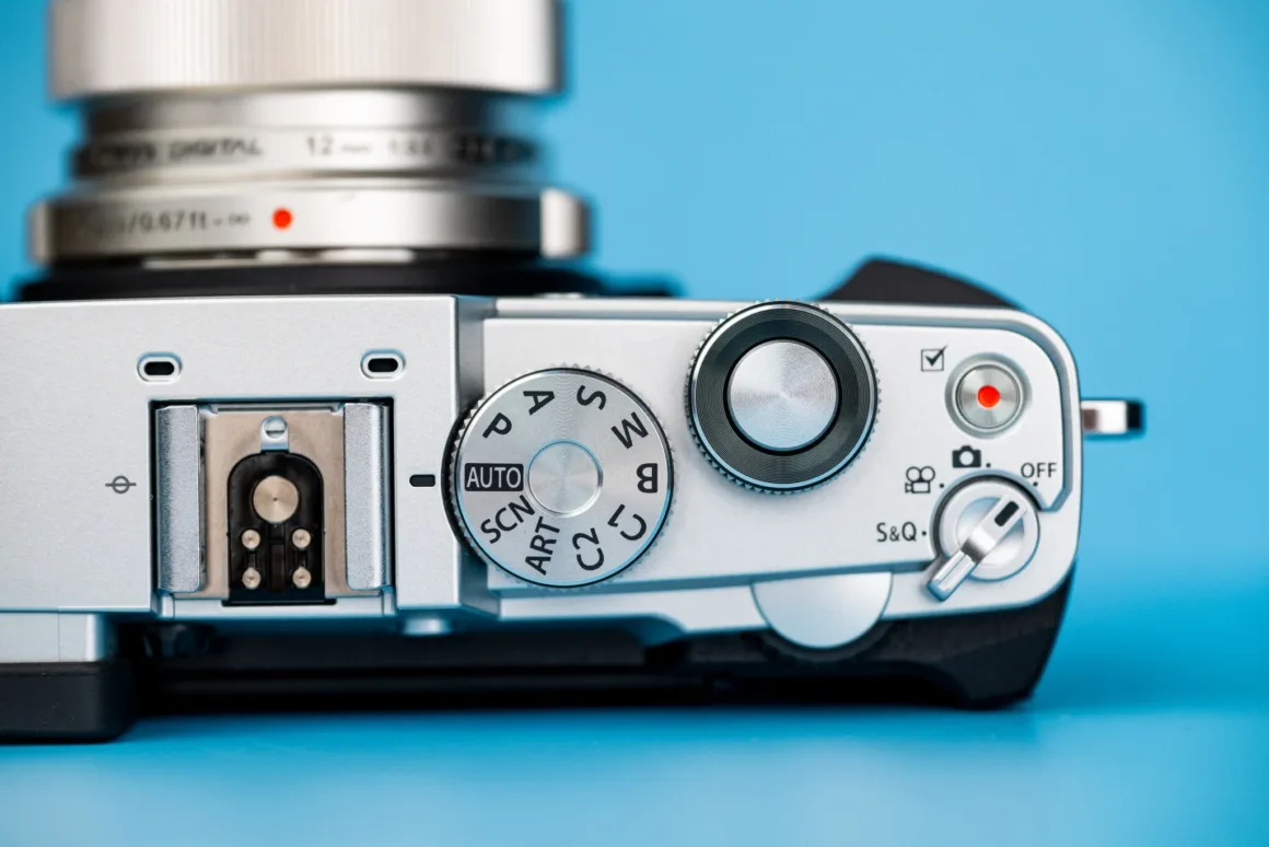 OM System PEN İncelemesi: Tanıdık Ama Son Derece Şık 22 A top-down view of a silver digital camera body with a lens, featuring a mode dial, shutter button, and power switch against a blue background.