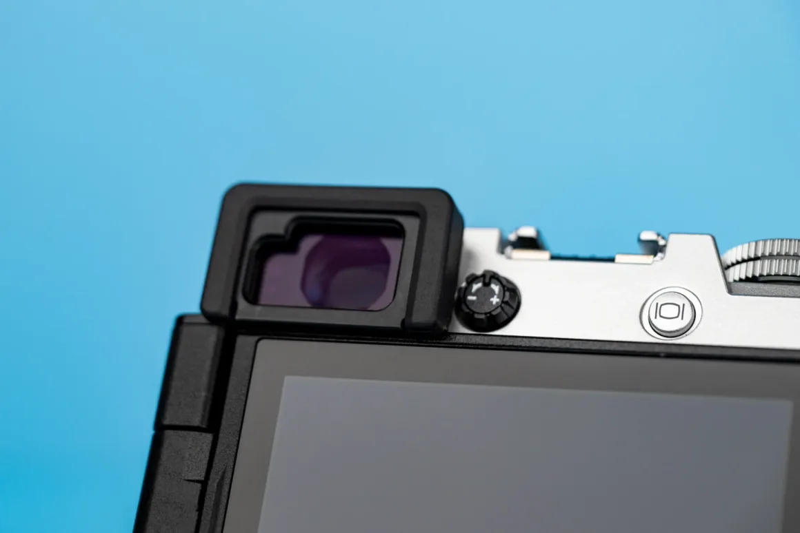 OM System PEN İncelemesi: Tanıdık Ama Son Derece Şık 24 A close-up view of the back of a silver and black digital camera, showing the electronic viewfinder, a diopter adjustment dial, and a display screen against a solid blue background.