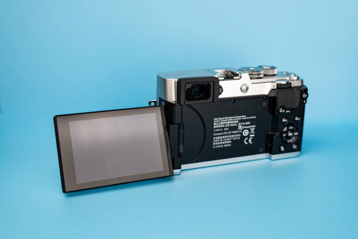 OM System PEN İncelemesi: Tanıdık Ama Son Derece Şık 25 A silver and black digital camera with its rear LCD screen flipped out to the left, set against a solid light blue background.