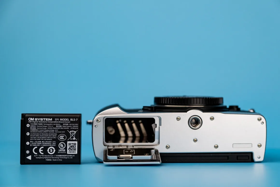 OM System PEN İncelemesi: Tanıdık Ama Son Derece Şık 26 An OM SYSTEM BLS-7 camera battery sits next to a silver camera with its battery compartment door open, set against a blue background.