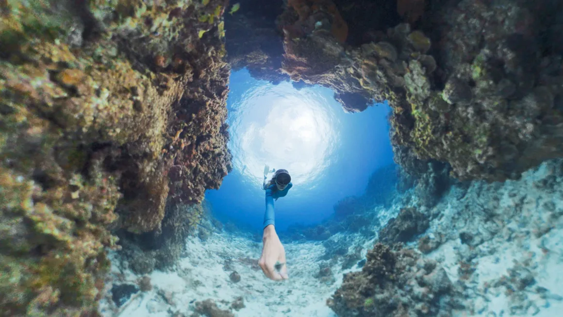 A freediver swims through a narrow underwater rock formation toward the bright surface, extending one arm toward the camera.
