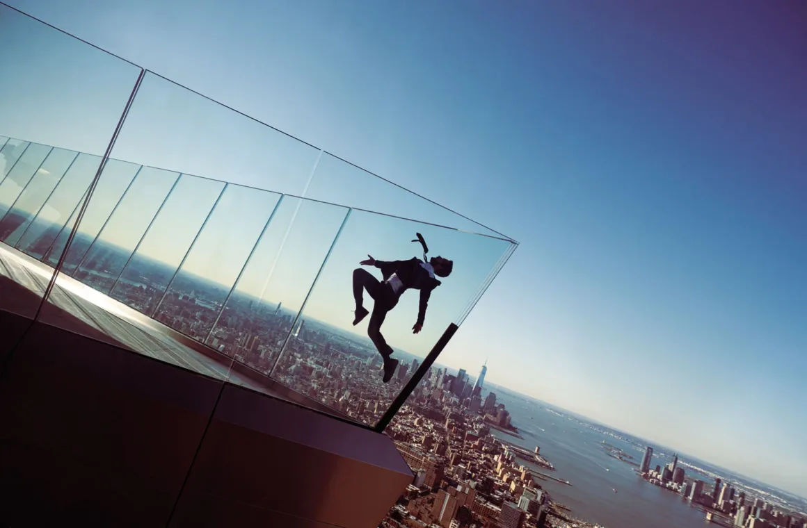 A person in a suit jumps in the air against a backdrop of a city skyline and a body of water, viewed from behind a glass barrier.