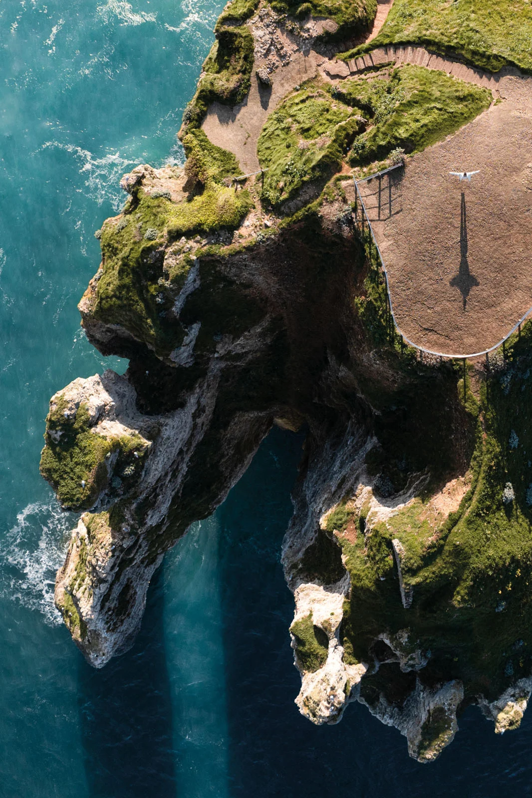 An aerial view of a person standing with arms outstretched on a grassy cliff edge overlooking the blue ocean.
