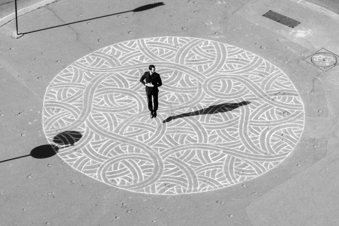 A man in a dark suit jumps in the center of a large, circular chalk drawing on an asphalt surface.