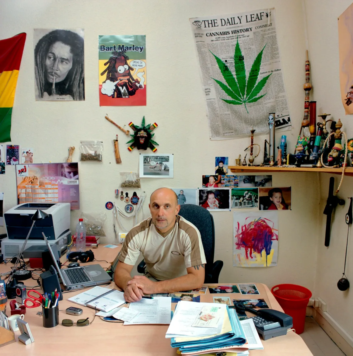 Dünyanın Dört Bir Yanından Bürokrat Portreleri 24 Bir adam, Bob Marley, Bart Simpson ve kenevir temalı görsellerle dekore edilmiş ofiste masasının başında oturuyor.
