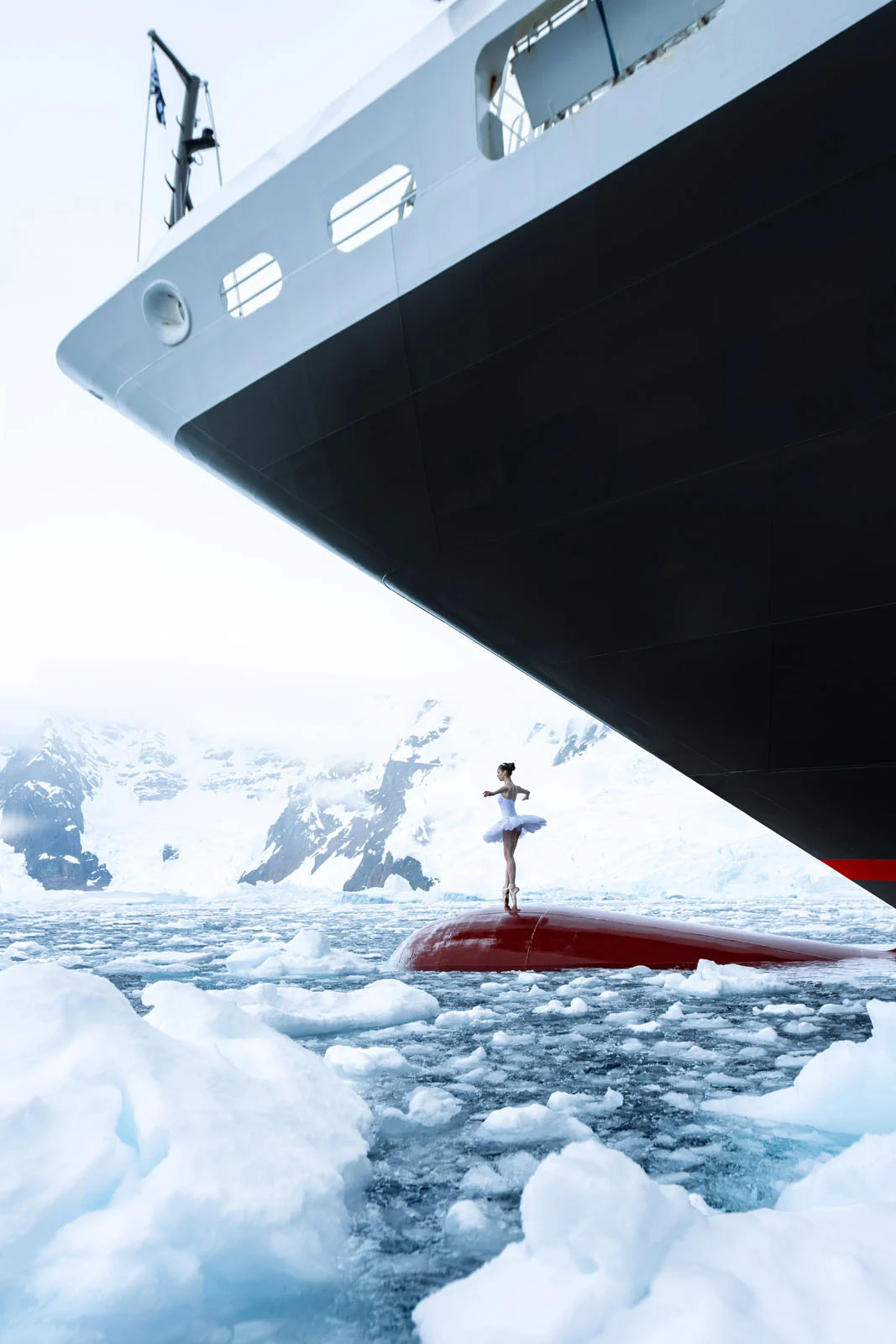 A ballerina in a white tutu stands on the red bulbous bow of a large ship surrounded by icy waters and snowy mountains.