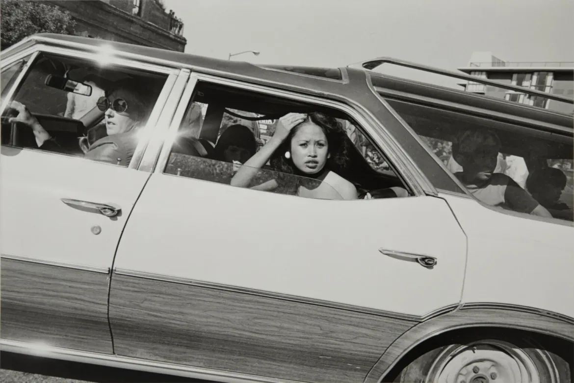 A black-and-white photograph shows a woman with dark hair looking out the window of a wood-paneled station wagon, with a driver wearing sunglasses and other passengers visible inside.