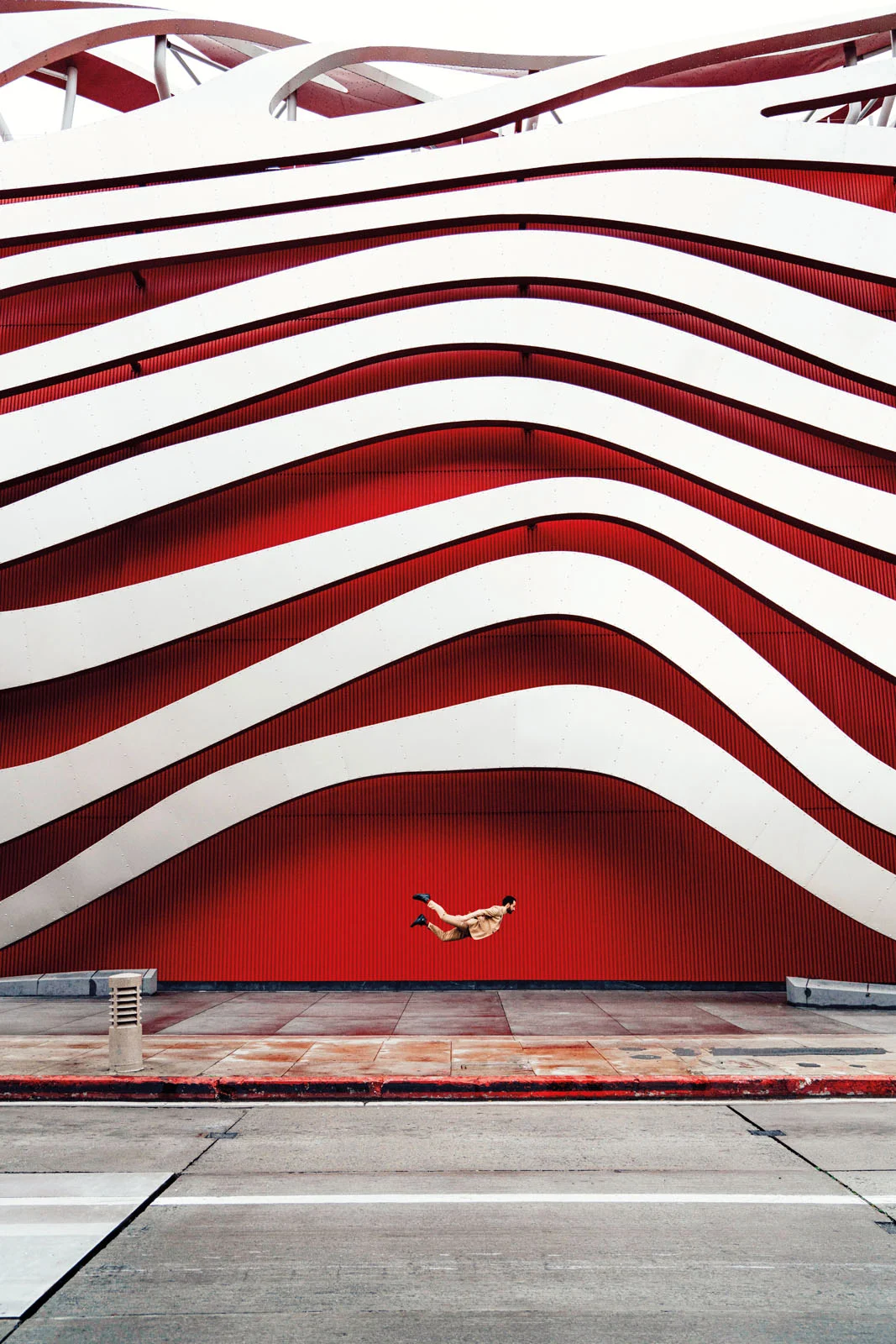 A person in a tan suit is captured mid-air in a horizontal jumping pose against a large wall featuring bold, wavy red and white stripes.