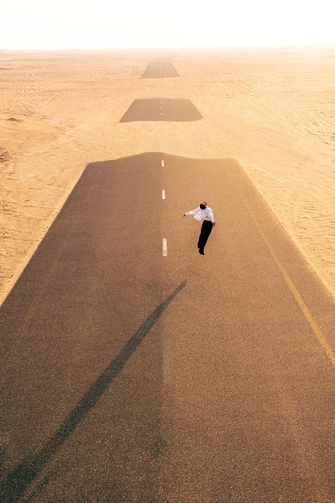 A man in a white shirt and dark trousers floats above a series of disconnected road segments surrounded by desert sand.