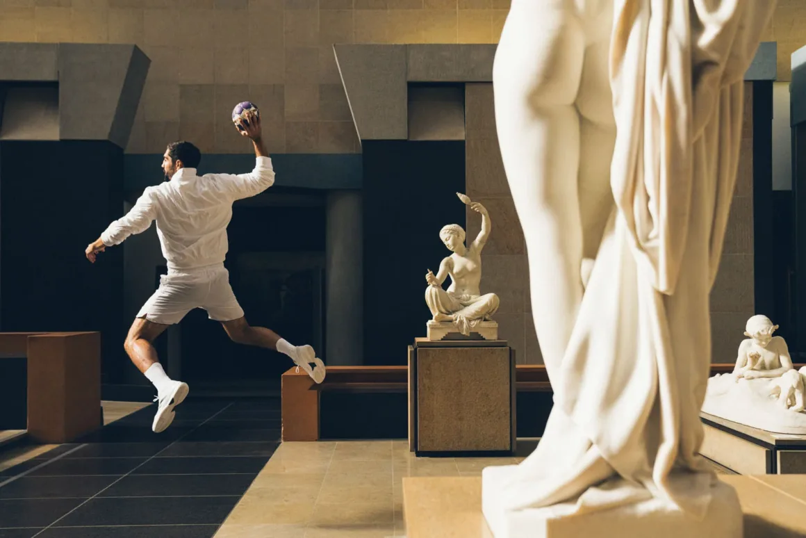 A man in white athletic clothing jumps in mid-air while holding a handball in a museum gallery filled with marble statues.