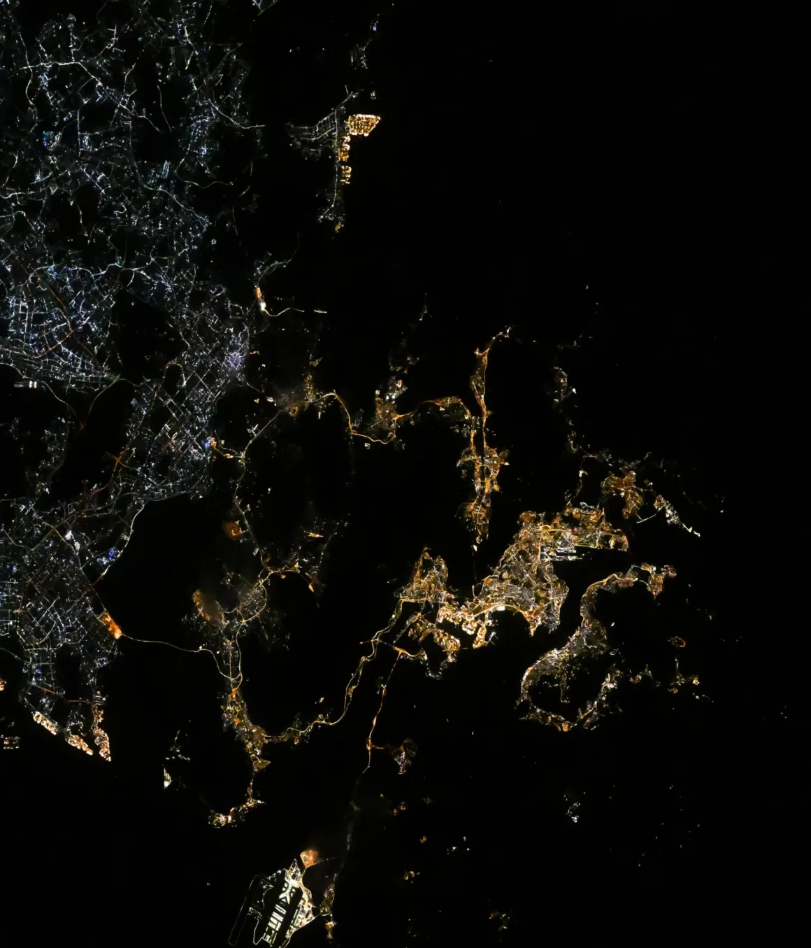 Astronotun Önce-Sonra Fotoğrafları Kentlerin Turuncudan Beyaz Işığa Geçişini Gösteriyor 32 A satellite view of a coastline at night, showing bright clusters of city lights against a dark landscape and water.