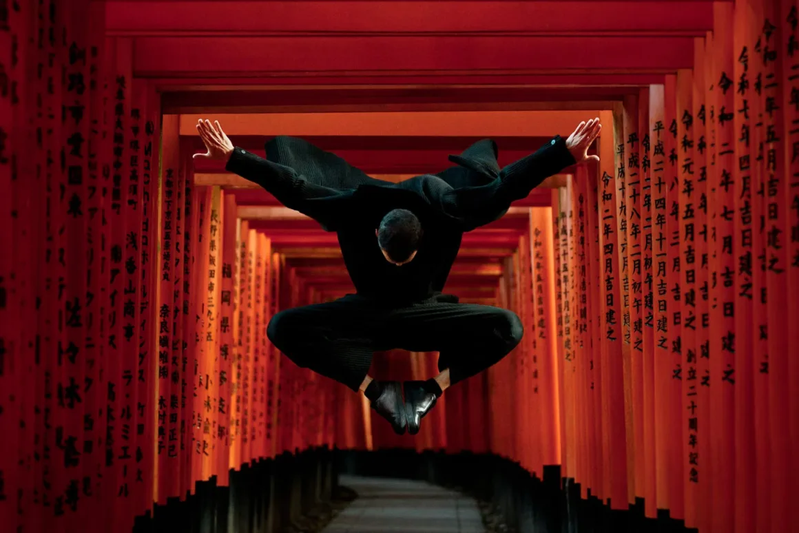 A person in an all-black outfit jumps mid-air with arms outstretched between two rows of bright red torii gates inscribed with Japanese characters.