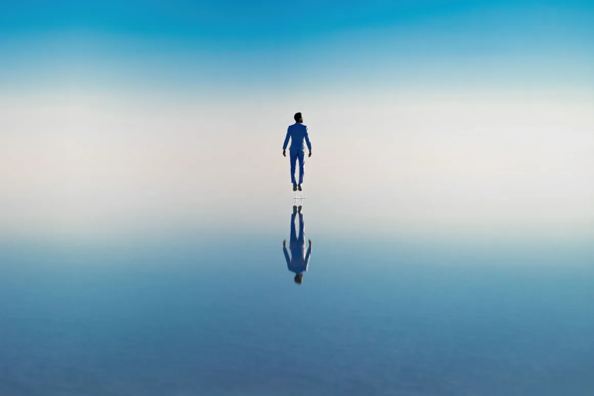 A person in a light blue suit floats above a calm, reflective surface that mirrors their figure against a clear blue sky.