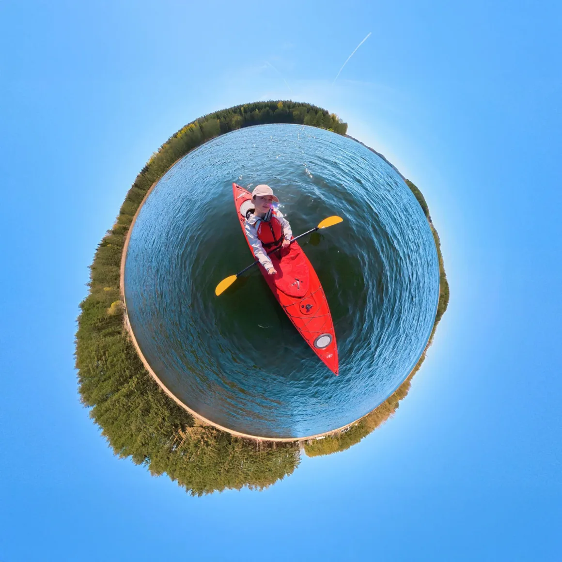 A person in a red kayak paddles on a lake, captured in a 360-degree spherical projection with a forest shoreline encircling the water.
