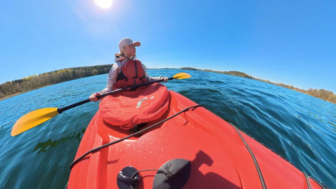A person wearing a life vest and a cap paddles a red kayak across a calm lake under a bright, clear blue sky.