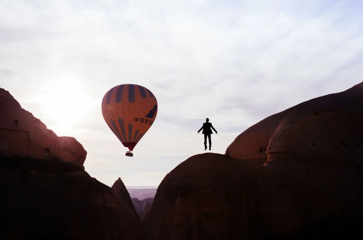 A silhouette of a person jumping in the air stands between two large rock formations, with a hot air balloon floating in the sky nearby.