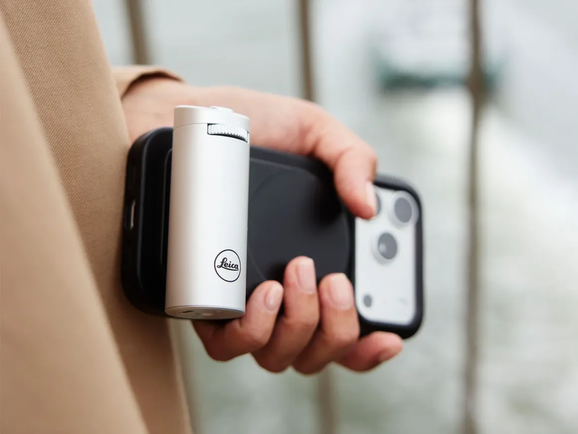 Leica Lux 2.0’a Steve McCurry İmzalı Yeni Görünüm Geldi 27 Bir kişi, arka kısmına gümüş renkli silindirik Leica markalı aksesuar takılmış bir akıllı telefon tutuyor.