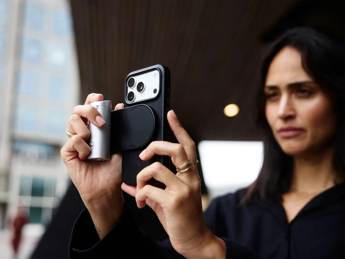 Leica Lux 2.0’a Steve McCurry İmzalı Yeni Görünüm Geldi 29 Bir kadın, yana doğru bakarken gümüş renkli silindirik kamera grip’i takılmış bir akıllı telefon tutuyor.