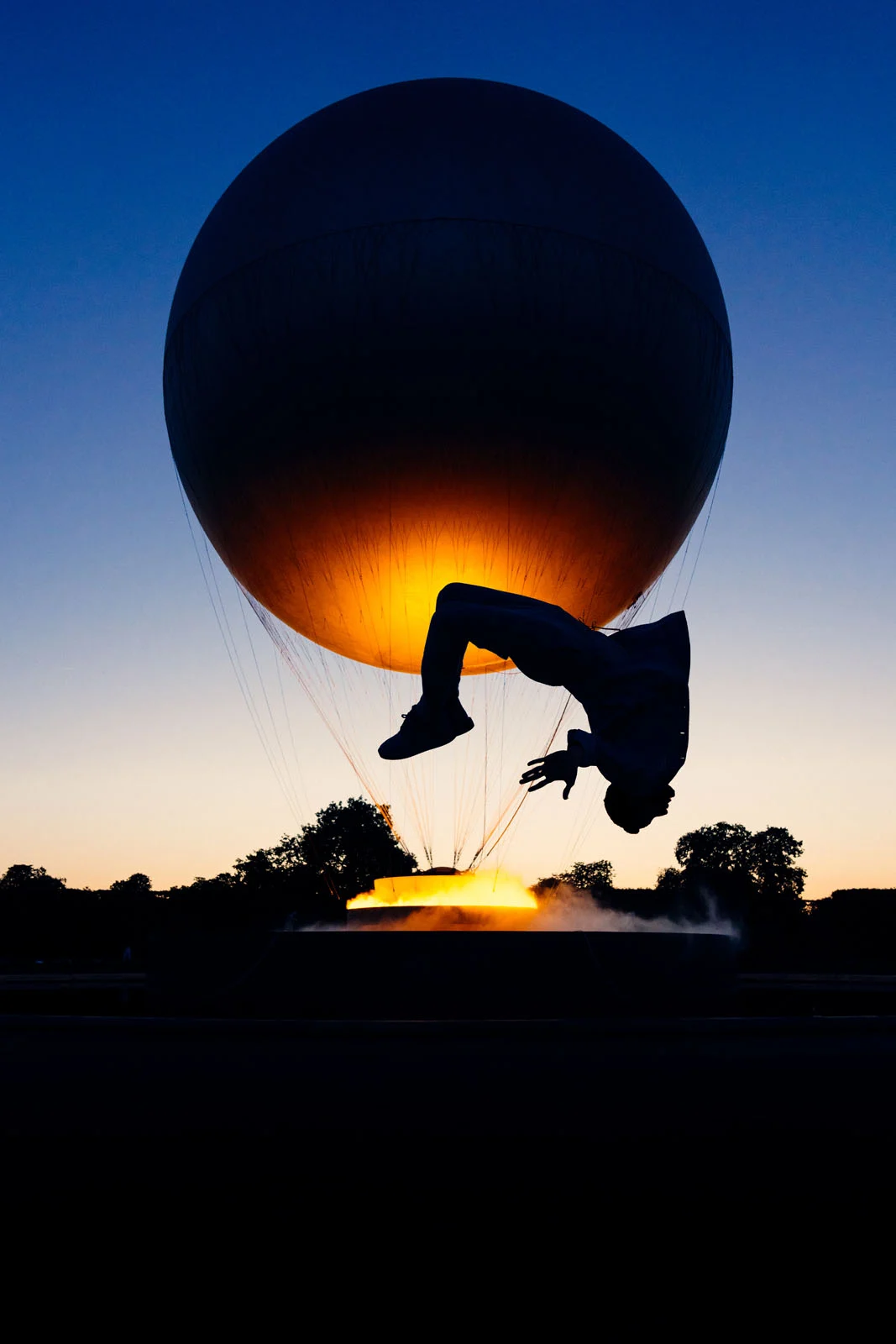 A silhouette of a person performing a backflip in front of a large, glowing, spherical structure at dusk.
