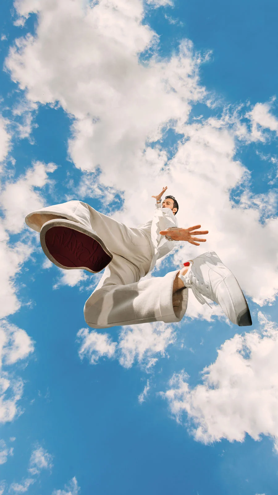 A man in a white outfit jumps against a bright blue sky filled with white clouds, captured from a low angle with his feet prominent in the foreground.
