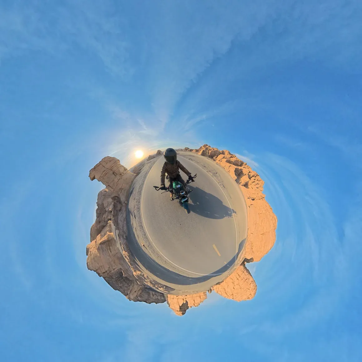 A 360-degree panoramic view shows a motorcyclist wearing a helmet and leather jacket riding on a desert road surrounded by rock formations under a bright sun.