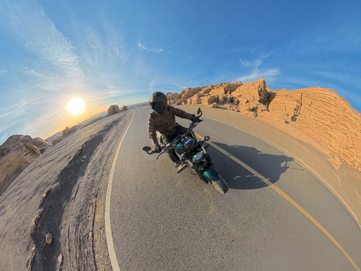 A motorcyclist wearing a helmet and brown jacket rides a motorcycle along a paved road through a desert landscape with rock formations under a sunny sky.