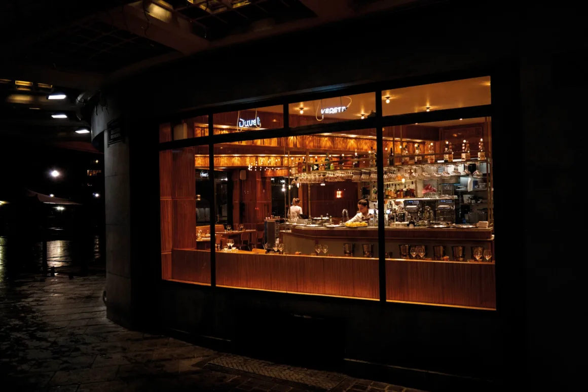 Nicolas Rachline’ın Tekinsiz Fotoğraflarında Modern Yaşamın Yalnızlığı ve Güzelliği 19 Gece büyük bir pencereden görülen, ahşap tezgâhın arkasında çalışan barmenin bulunduğu aydınlık bar.