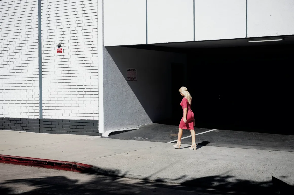 Nicolas Rachline’ın Tekinsiz Fotoğraflarında Modern Yaşamın Yalnızlığı ve Güzelliği 21 Pembe elbiseli ve yüksek topuklu sarışın bir kadın güneşli bir günde otopark girişinin önünden geçiyor.