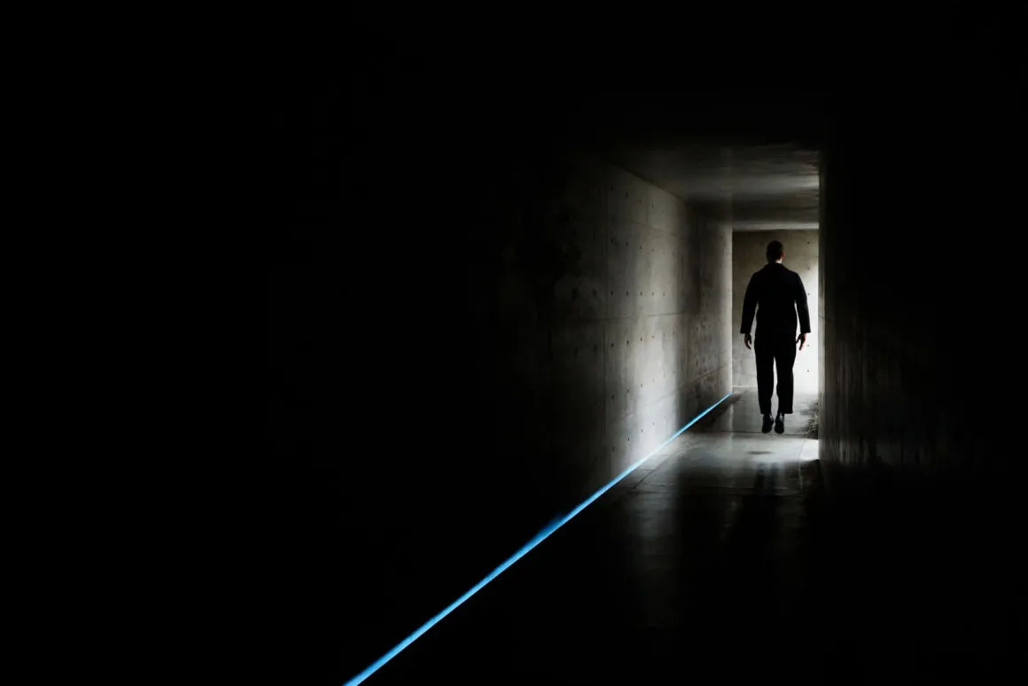 A person in a dark suit levitates slightly above the floor in a dimly lit, narrow concrete corridor with a glowing blue floor light.