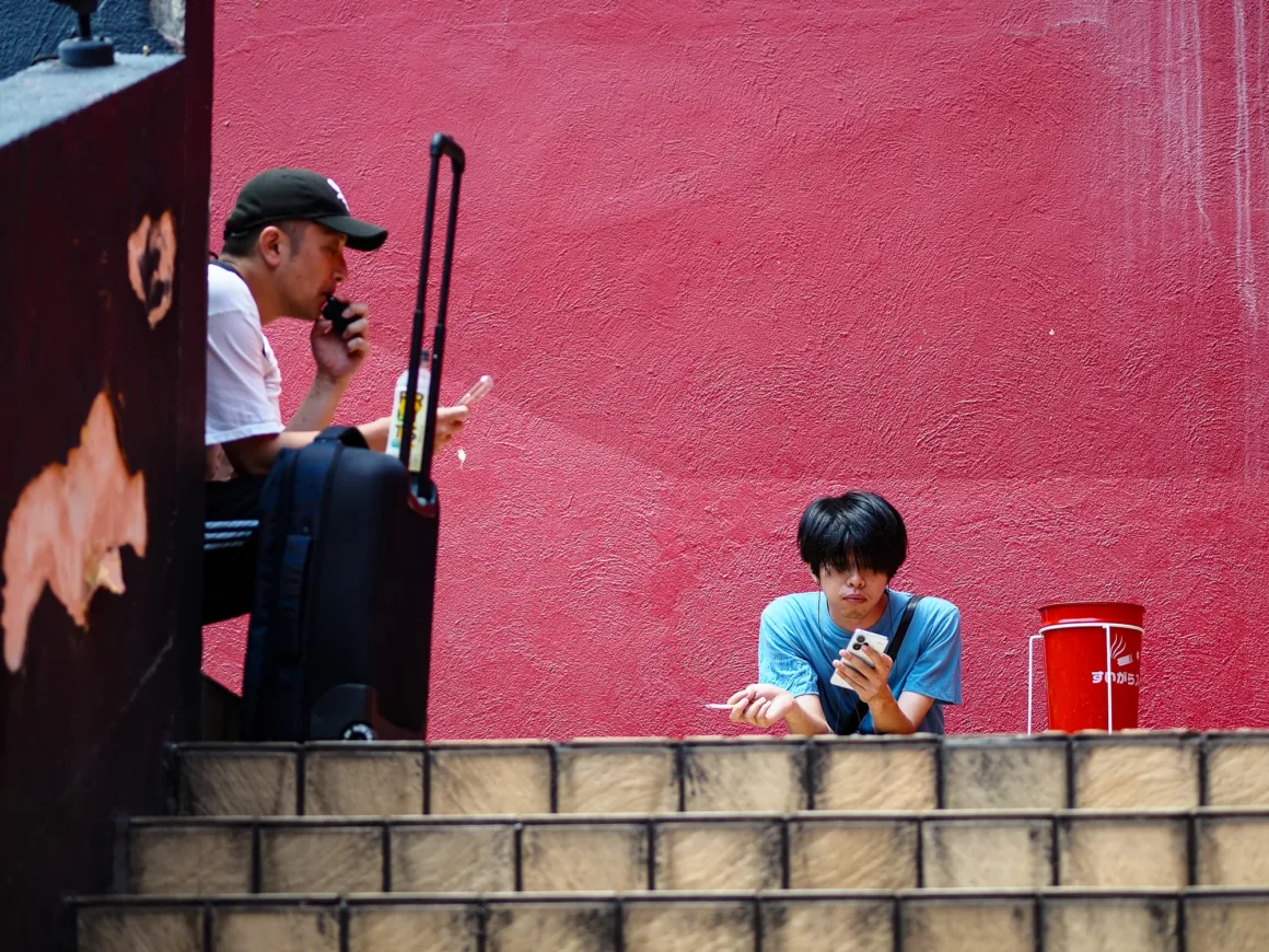 OM System PEN İncelemesi: Tanıdık Ama Son Derece Şık 47 Two men sit on outdoor stairs against a bright red wall, one using a vape and the other holding a smartphone and a cigarette.