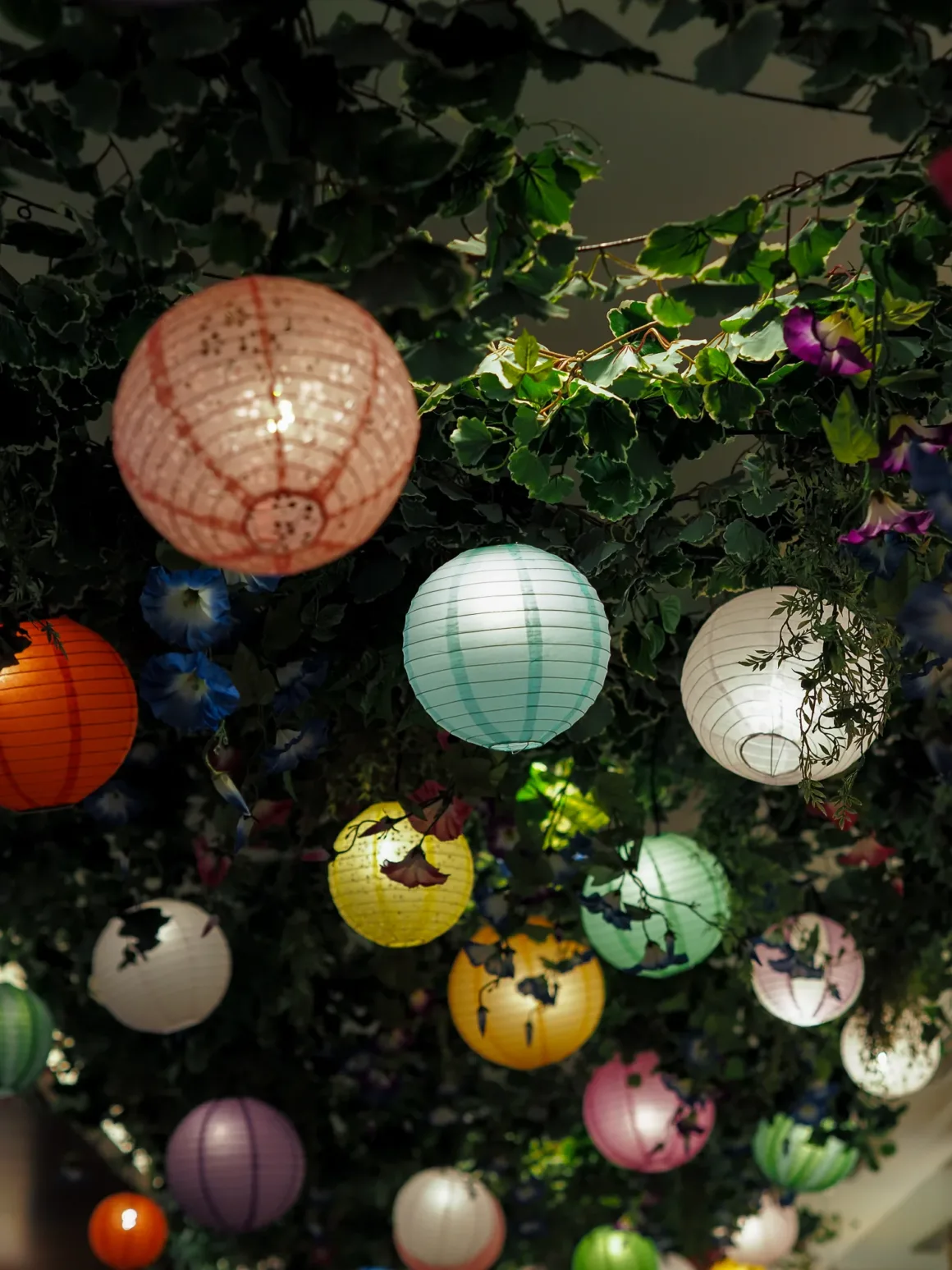 OM System PEN İncelemesi: Tanıdık Ama Son Derece Şık 46 A collection of colorful, glowing paper lanterns hangs from a ceiling covered in lush green foliage and blue flowers.