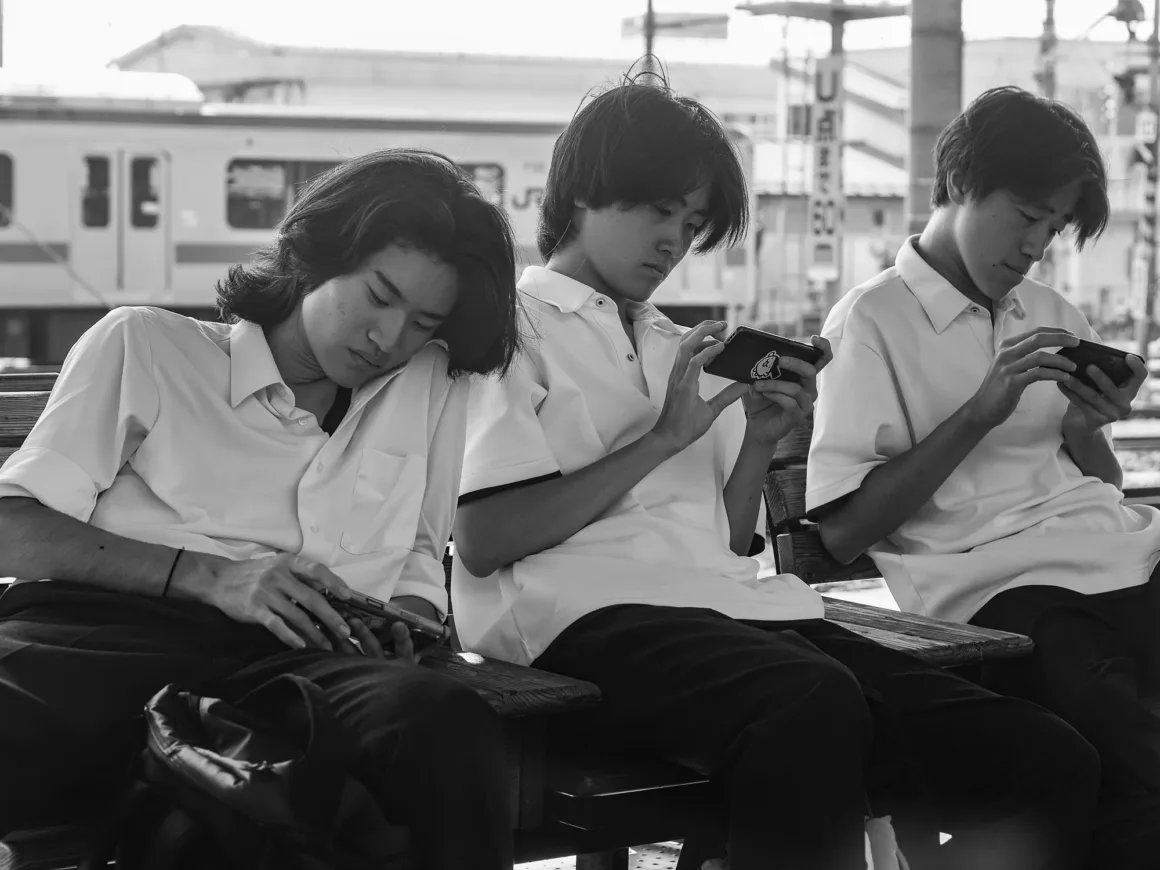 OM System PEN İncelemesi: Tanıdık Ama Son Derece Şık 32 Three young men in white collared shirts sit on a wooden bench at a train station, each looking down at a smartphone.